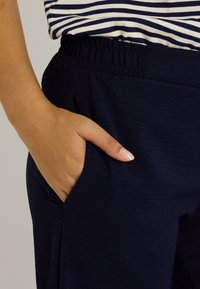 Pantalon bleu marine en tissu texturé, avec une taille élastique et des poches latérales. Main placée dans une poche, portant un haut rayé.