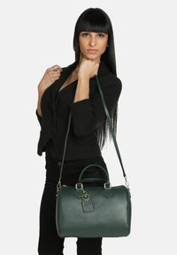 Sac à main en cuir vert avec une forme structurée, doté de poignées supérieures et d'une bandoulière amovible, agrémenté de quincaillerie dorée et d'une étiquette de bagage.