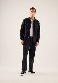 Veste polaire noire avec deux poches poitrine et fermeture à boutons, t-shirt blanc, pantalon noir et baskets noires avec des accents blancs.