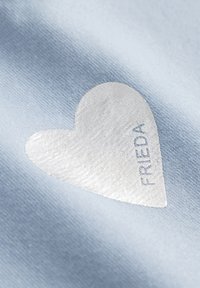 Lichtblauwe stof heeft een zachte, textuurachtige witte hart met het woord "FRIEDA" subtiel in reliëf gedrukt.