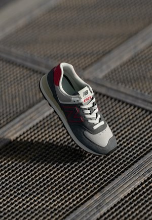 Baskets New Balance 574 gris et rouges avec lacets blancs, posées sur une surface métallique texturée, inclinées pour montrer les vues latérale et supérieure.