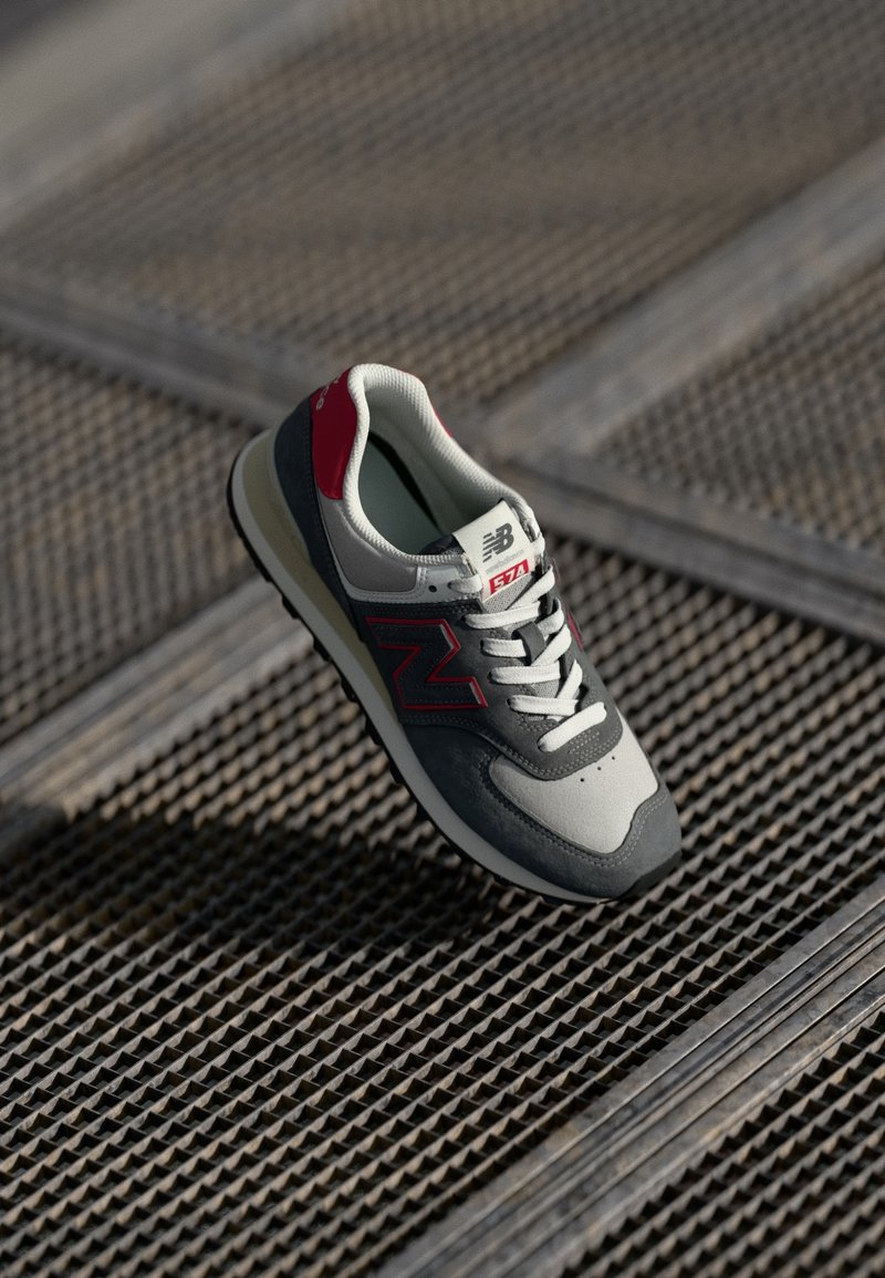 Baskets New Balance 574 gris et rouges avec lacets blancs, posées sur une surface métallique texturée, inclinées pour montrer les vues latérale et supérieure.