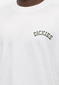 Λευκό βαμβακερό μπλουζάκι με καμπυλωτό πράσινο λογότυπο "DICKIES" στην άνω αριστερή περιοχή του στήθους. Απαλή υφή με κλασικό σχεδιασμό εφαρμογής.