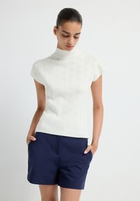 White knit sweater with a high neck and short sleeves, featuring a raised leaf pattern. Paired with navy tailored shorts.
