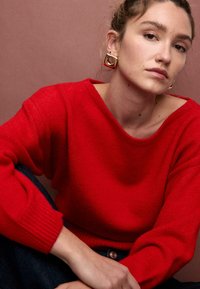 Roter Strickpullover mit weitem Boot-Ausschnitt und gerippten Bündchen, kombiniert mit dunkelblauer Jeans. Modell trägt auffällige, geometrische goldene Ohrringe.