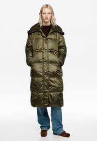 Padded long coat in olive green with a textured dark collar, featuring large pockets and a button closure. Grey-blue wide-leg jeans worn underneath.