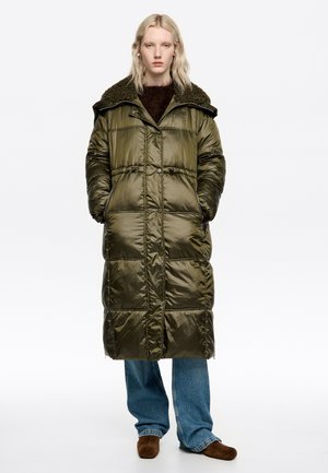 Padded long coat in olive green with a textured dark collar, featuring large pockets and a button closure. Grey-blue wide-leg jeans worn underneath.