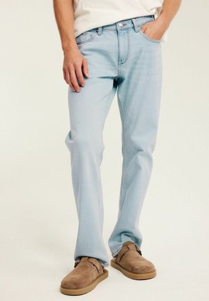 Mann trägt hellblaue gerade geschnittene Jeans mit braunen Wildleder-Slippern, steht mit einer Hand in der Tasche.