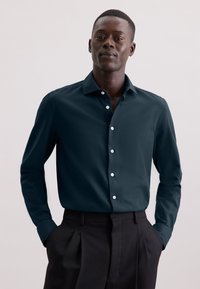 Camicia a maniche lunghe in verde scuro con colletto a punta, caratterizzata da bottoni bianchi e una texture liscia, indossata infilata in pantaloni neri.
