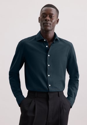 Donkerblauwe langarmshirt met een puntige kraag, voorzien van witte knopen en een gladde textuur, gedragen in een zwarte broek.
