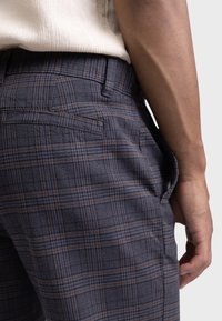 Dark plaid trousers featuring a textured fabric, with thin blue, brown, and gray lines. Back pocket detail visible, with a tailored fit.