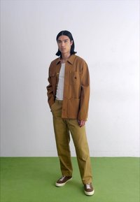 Giacca in tela marrone con tasche frontali e dettagli in oro, abbinata a pantaloni verdi oliva e sneakers marroni con lacci bianchi.