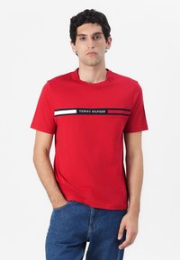 Červené bavlněné tričko s kulatým výstřihem a krátkým rukávem, s černobílým horizontálním pruhovaným designem a logem "TOMMY HILFIGER".