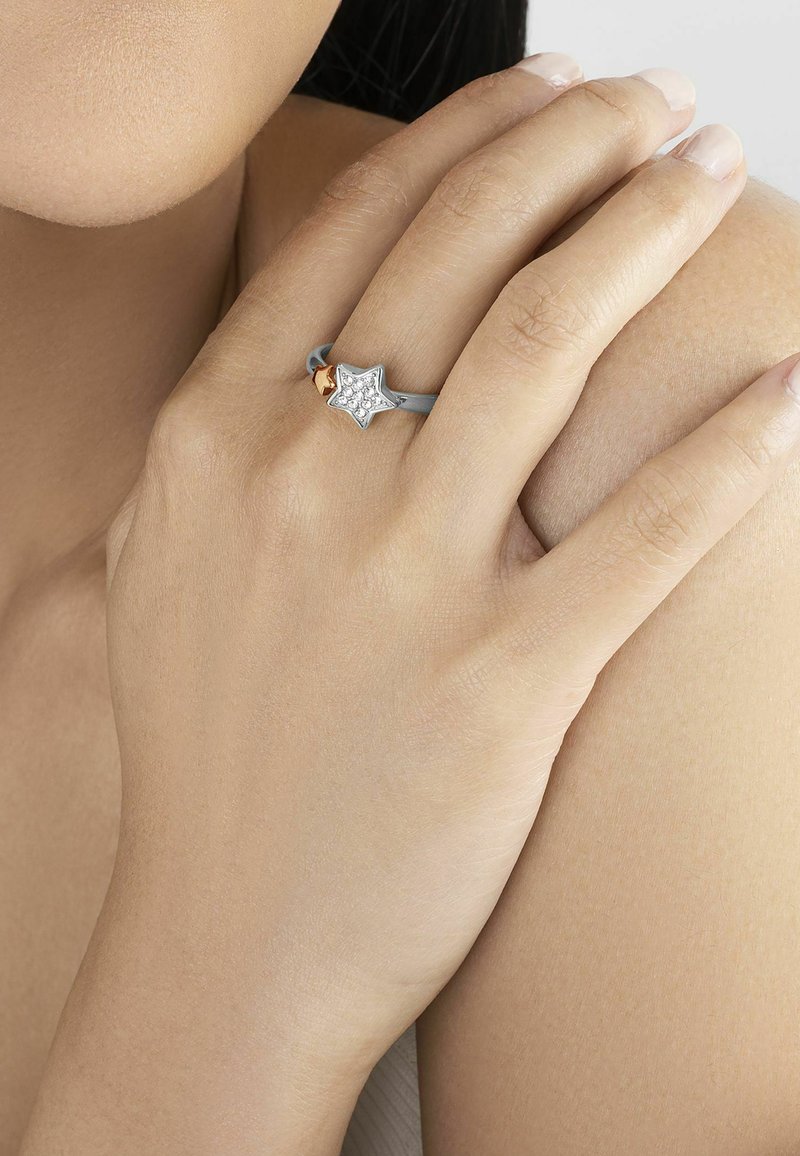 Söljfärgad stjärnformad ring med en kluster av klara ädelstenar, accentuerad av ett litet guld-element, bärs på en hand utan nagellack.