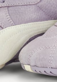 Lavender sneaker made of textured leather with a suede white stripe. Features visible stitching and a rubber sole with a curved design.