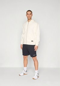 Krémová mikina na zip s kapsami vpředu, doplněná tmavými šortkami. Modelka má na nohou bílé tenisky s světle hnědými podrážkami, stojící na jednobarevném pozadí.