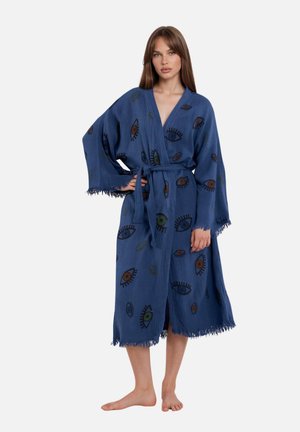 Vrouw draagt een blauwe kamerjas met oogmotieven, gestrikt in de taille, met wijde mouwen en franjeranden, blootvoets staand op een witte achtergrond.