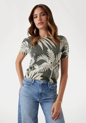 Il top lavorato a maglia presenta una base verde scuro con un motivo di foglie di palma color crema. Maniche corte e texture a coste. Abbinato a jeans azzurri.