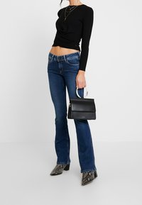 Svart långärmad topp med en vriden design, blå utsvängda jeans, grå ankelboots med ormskinnsmönster och en liten svart handväska med ett cirkulärt handtag.