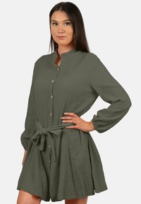 Abito camicia verde oliva scuro realizzato in tessuto texturizzato. Presenta un colletto, chiusura con bottoni e dettaglio cintura da annodare, con maniche lunghe e orlo svasato.