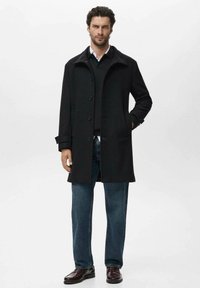 Abrigo de lana negro con un cuello estructurado, cierre de botones, bolsillos laterales, llevado sobre un suéter negro y unos vaqueros azules, combinado con zapatos oscuros.