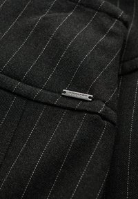 Schwarzer Stoff mit Nadelstreifen und strukturiertem Finish, mit einem silbernen "Superdry"-Etikett auf einer Tasche. Klare Linien und strukturierte Designdetails.