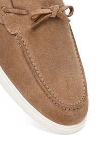 Zapato tipo mocasín de ante marrón con detalles cosidos, un lazo en la parte superior y una suela de goma blanca.