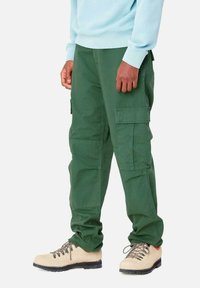 Groene cargo shorts met meerdere zijzakken, gemaakt van duurzaam materiaal. Gecombineerd met beige laarzen met zwarte zolen en veters.