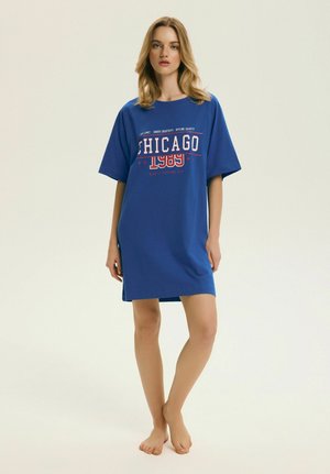 Mladá žena stojaca bosá, oblečená v voľné modré šaty s nápisom „CHICAGO 1989“ na prednej strane na jednoliatom pozadí.