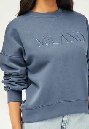 Femme portant un sweat-shirt bleu avec "MILANO" brodé sur la poitrine et un jean bleu clair.