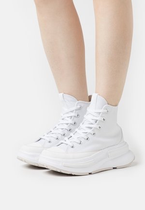 Baskets montantes blanches avec semelles épaisses portées sur des jambes nues, vues de face en angle sur un fond blanc uni.