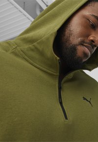 Olivengrøn hoodie med lynlåskrave, blød tekstur og sort Puma-logo. Har en hætte og et afslappet fit design.