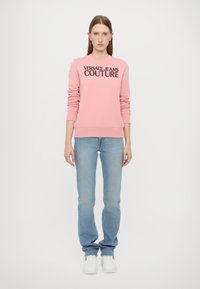 Rozā sporta krekls ar "VERSACE JEANS COUTURE" logotipu, kombinēts ar gaiši ziliem džinsiem un baltām kedām; ribotie piedurkņu un apakšmalas gali.