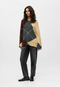 Oversized v-neck knit sweater featuring a geometric patchwork design in brown, gray, and beige, with soft texture. Paired with dark trousers.