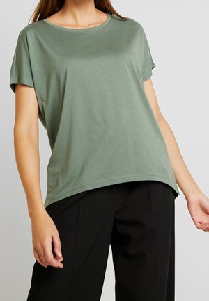 Frau trägt ein lockeres grünes kurzärmeliges Oberteil und schwarze Hose, steht mit entspannten Armen vor einem schlichten hellen Hintergrund.