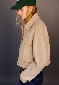 Beige cropped wool jacket with a collar, front pockets, and button details, paired with blue jeans and a green cap.