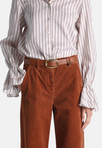 Chemise blanche boutonnée avec des rayures rouges et des poignets froufroutants, associée à un pantalon en velours côtelé couleur rouille et une ceinture marron avec une boucle dorée.
