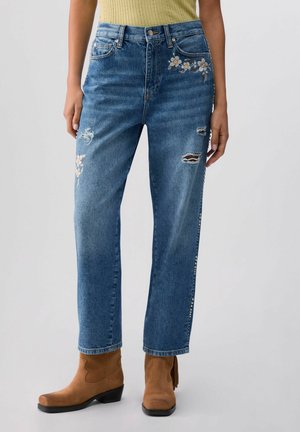 Kobieta ubrana w niebieskie podarte dżinsy z kwiatowym haftem i ćwiekami po bokach, w połączeniu z beżowymi botkami za kostkę oraz żółtym prążkowanym topem.