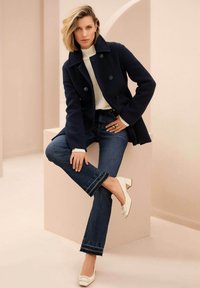 Navy peacoat with double-breasted front, white knit sweater, dark denim jeans, and white block-heeled shoes. Smooth textures and clean lines.