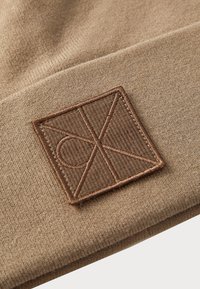 Pruun kudumine müts, mis on valmistatud pehmest materjalist ja millel on tekstuuriga ruudukujuline plaaster tumedama pruuniga kirja logoga.
