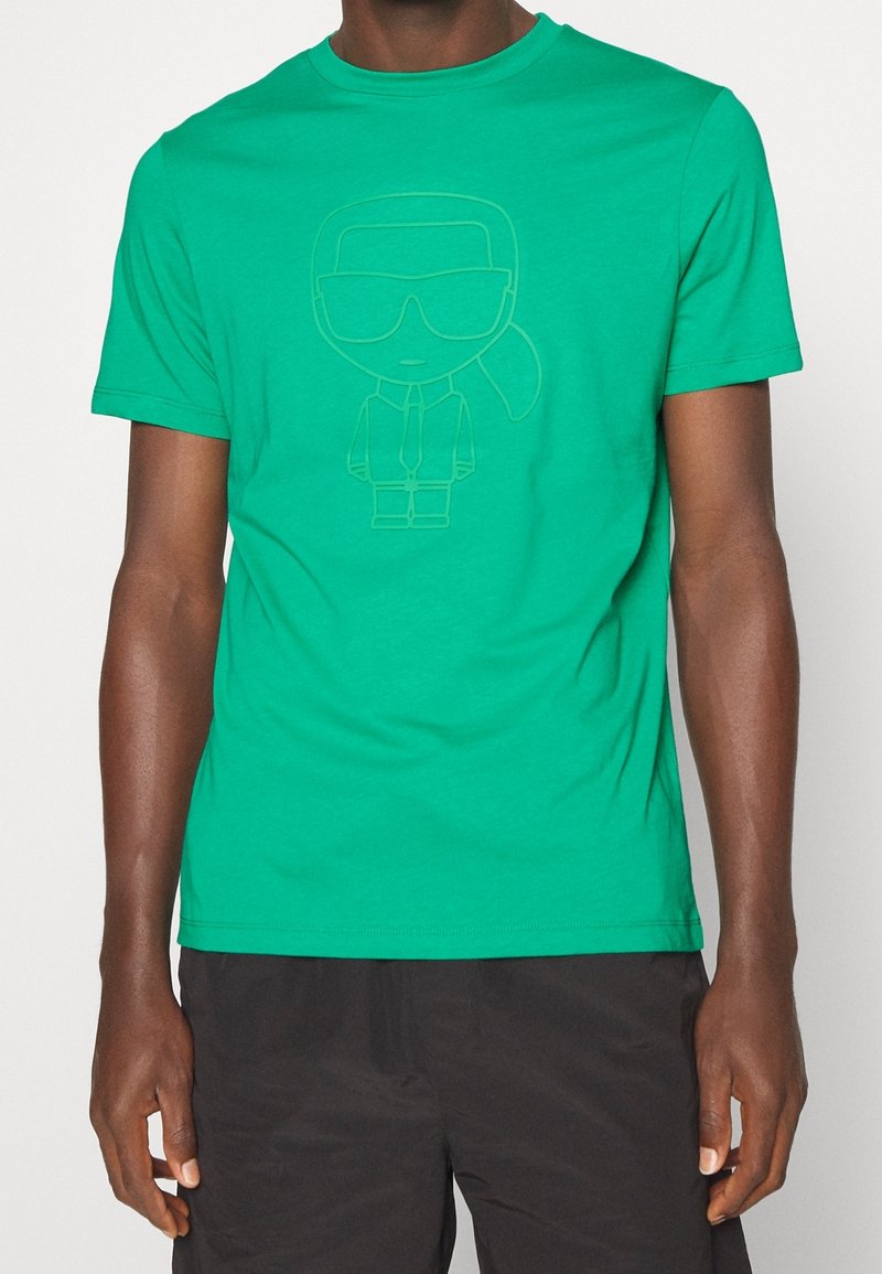 Zielona bawełniana koszulka z krótkim rękawem, ozdobiona minimalistyczną postacią w garniturze i okularach przeciwsłonecznych. Okrągły dekolt i gładka faktura.