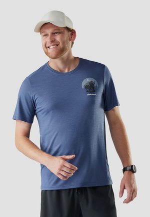 Lächelnder Mann mit weißer Mütze, blauem T-Shirt mit Bärengrafik und Schriftzug "Smartwool", schwarzen Shorts und einer Armbanduhr am linken Handgelenk.