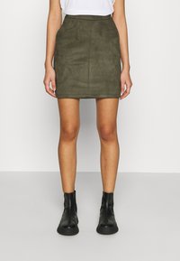 Kvinna klädd i en olivgrön minikjol, vit skjorta instoppad, och svarta stövletter, stående mot en enkel ljus bakgrund.