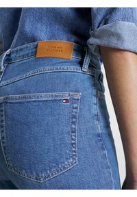 Ljusblå denimjeans med en brun läderlogotyp, med en liten broderad flagga på bakfickan. Klassisk fem-fickdesign.