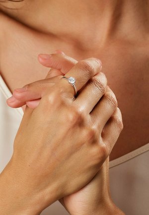 Silver ring with a round, clear gemstone, featuring a simple band design. Hand clasped with natural skin texture, indoors, neutral background.