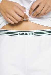 Witte katoenen ondergoed met een groen-wit gestreepte elastische tailleband, voorzien van de opdruk "LACOSTE" in het groen. Glad oppervlak, aansluitend ontwerp.