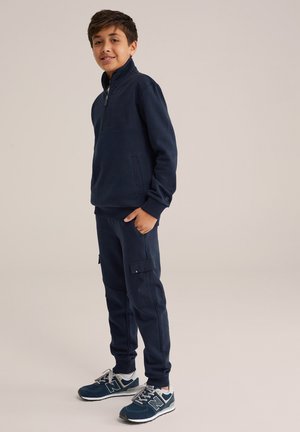 Marine blauwe fleece zip-up jas met een hoge kraag, gecombineerd met bijpassende cargo broeken met zijzakken en elastische boorden. Gestyled met sportieve sneakers.