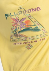 Yellow sweatshirt featuring a triangle graphic with palm trees, sun, and mountains, along with “BILLABONG” and “ESTABLISHED IN 1973” text.