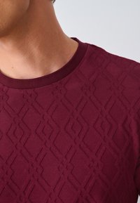 Burgundy sweater featuring a textured diamond pattern, round neckline, and ribbed collar. Smooth fabric with a slight sheen.