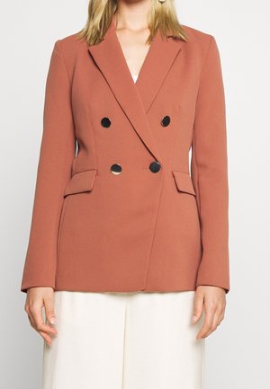 Vue recadrée d’une femme portant un blazer croisé couleur rouille avec des boutons noirs et un pantalon blanc.